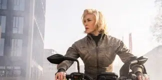 Woman in a brown jacket sits on a black motorcycle in an urban area, looking to the side with a helmet resting on the right handlebar.