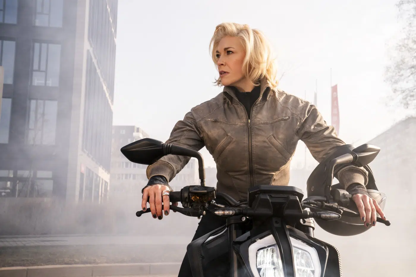 Woman in a brown jacket sits on a black motorcycle in an urban area, looking to the side with a helmet resting on the right handlebar.