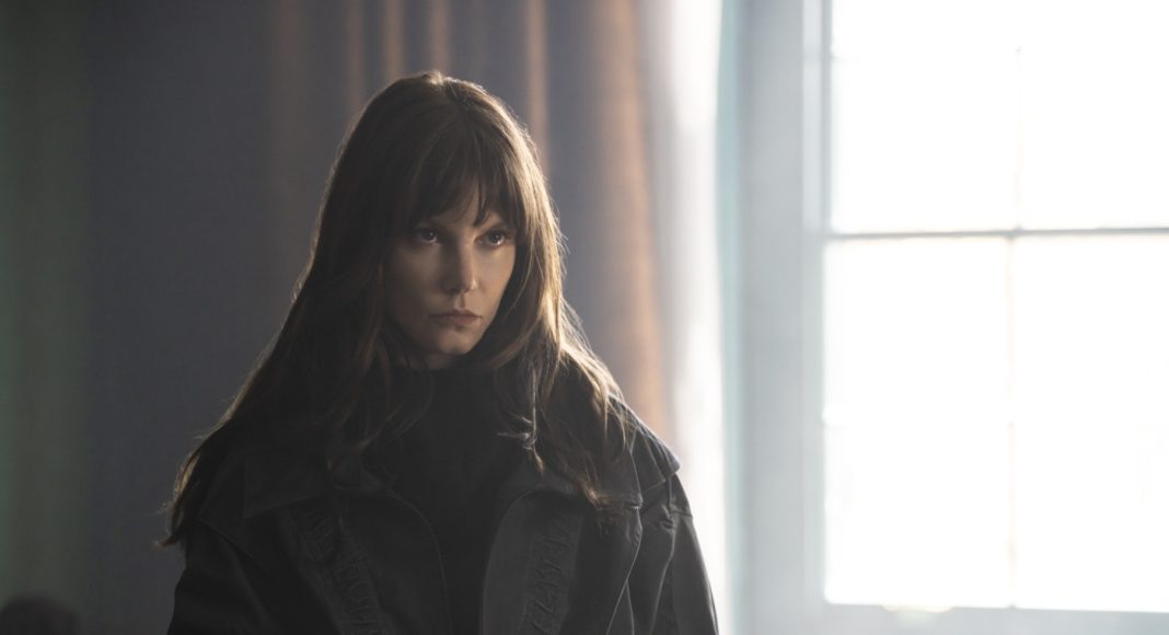 Young woman with long brown hair wearing a black coat stands in a dim room, gaze focused off to the side by a bright window.