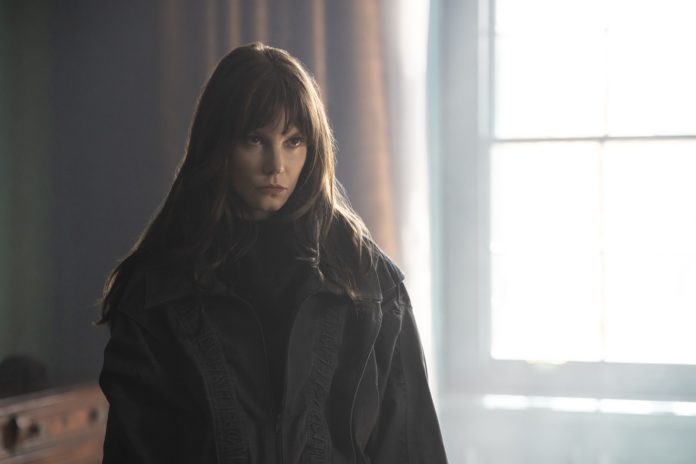 Young woman with long brown hair wearing a black coat stands in a dim room, gaze focused off to the side by a bright window.