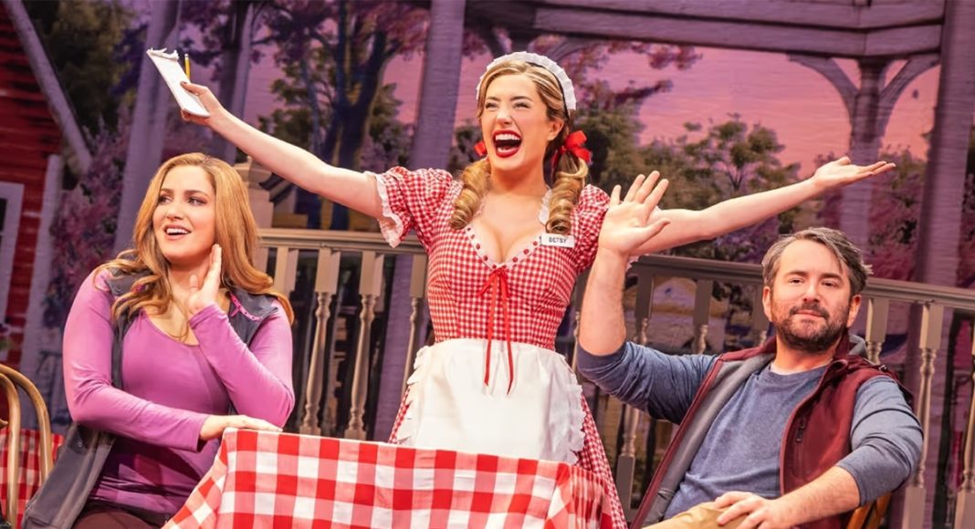Three performers on a stage: a smiling waitress in a red checkered dress raises her arms while two others watch and smile at a table with a red‑and‑white tablecloth.