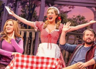 Three performers on a stage: a smiling waitress in a red checkered dress raises her arms while two others watch and smile at a table with a red‑and‑white tablecloth.