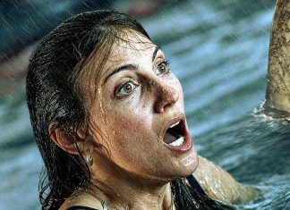 Woman in the ocean with wet hair, mouth open in alarm as a shark fin rises nearby.
