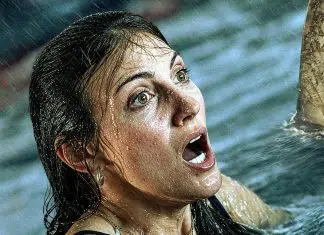 Woman in the ocean with wet hair, mouth open in alarm as a shark fin rises nearby.