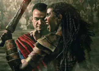 Two armored fighters collide in a dusty arena, one raising a bloodstained weapon as the crowd watches.