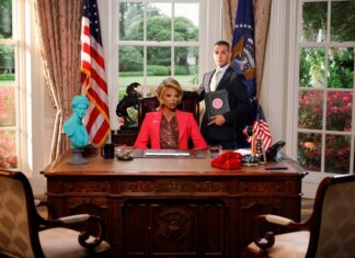 Woman in a red blazer sits at an ornate desk while a man in a suit stands beside her holding a binder in a formal office setting with flags in the background.