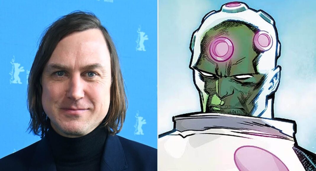 superman Left: man with shoulder-length hair in a dark blazer and black turtleneck against a blue backdrop; right: comic-style green-skinned figure with a pink brain-implant device.