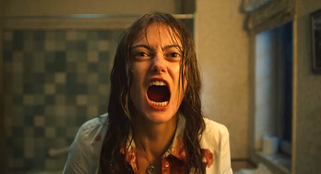 Woman with wet hair, mouth wide open in a scream, bloodstains on a white shirt, standing in a dim, tiled bathroom.