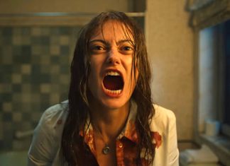 Woman with wet hair, mouth wide open in a scream, bloodstains on a white shirt, standing in a dim, tiled bathroom.