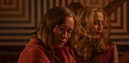 Two women sit side by side in a warm, dim room; the foreground woman wears glasses and an orange sweater.|Two women seated indoors with soft amber lighting, one in the foreground wearing glasses and a rust-colored sweater, looking down.|Contemplative scene of two women in a warmly lit room, with a geometric-patterned wall in the background.|Foreground: woman with glasses in an orange knit sweater; behind her, a blonde woman with curls in a red-toned setting.|Two thoughtful women in a cozy, amber-lit space, one in the foreground and the other slightly behind.