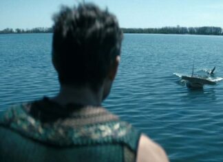 Back of a person with short hair and a patterned scarf stands at a blue lake, watching a small boat skimming the water ahead.