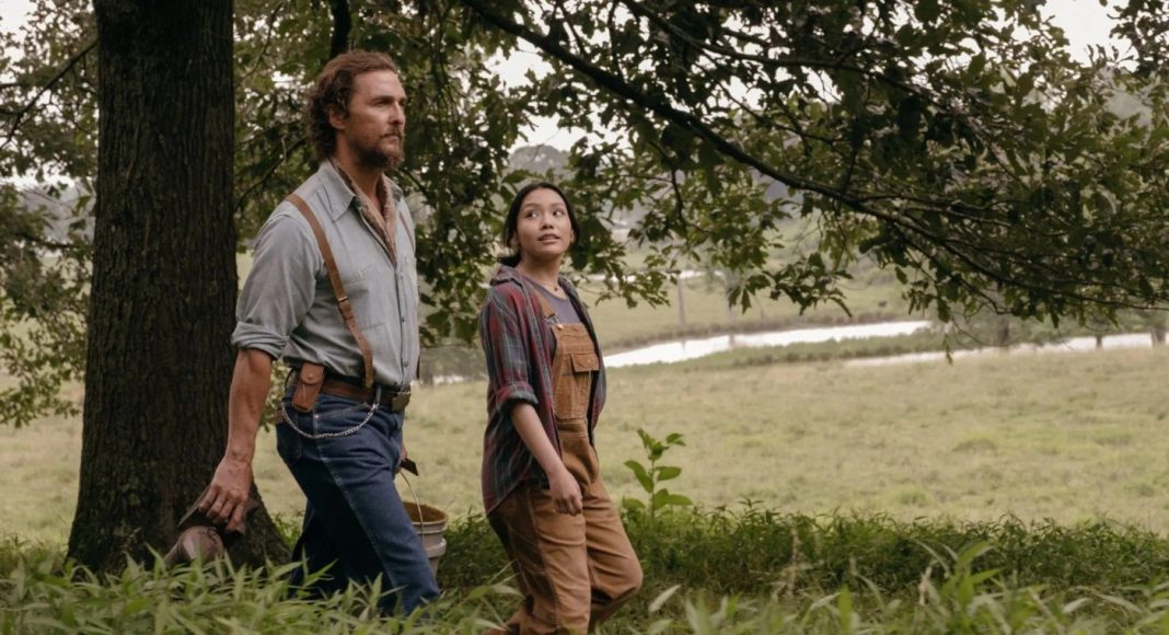 Two adults walking through a grassy rural area beside a tree, the man in a blue shirt with suspenders and the woman in overalls and a plaid shirt.
