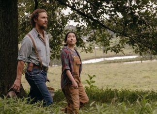 Two adults walking through a grassy rural area beside a tree, the man in a blue shirt with suspenders and the woman in overalls and a plaid shirt.