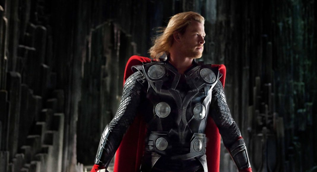 Thor in black armor and red cape stands in a dark cavernous setting, gripping Mjolnir in his left hand.