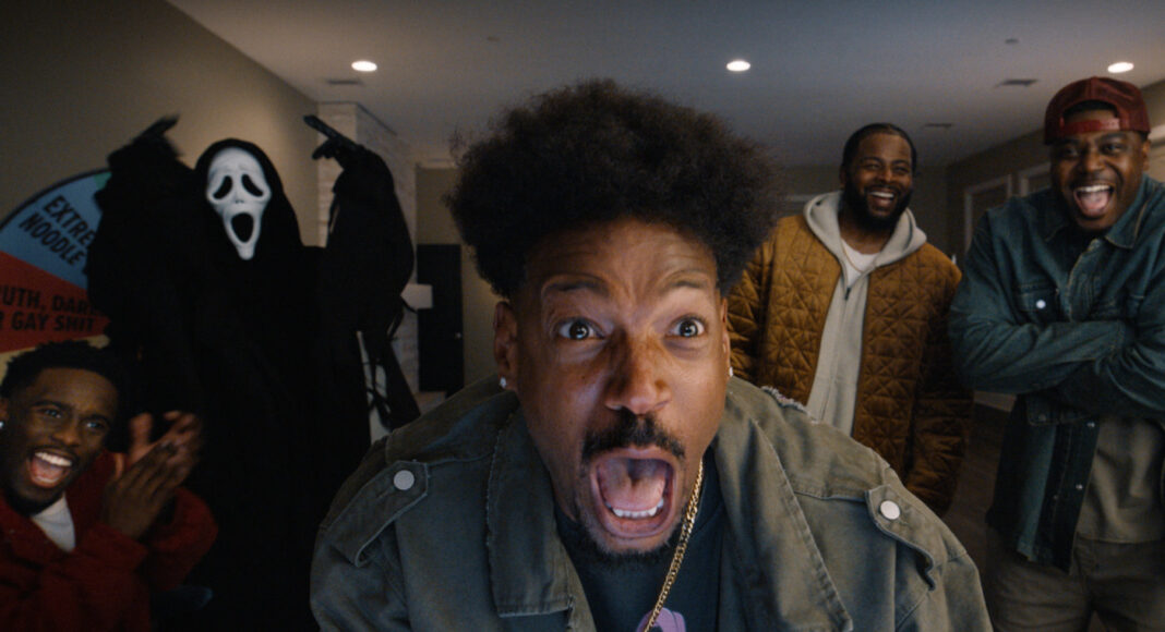 todo mundo em panico Close-up of a man screaming at the camera, surrounded by friends laughing indoors; a Ghostface-costumed figure stands in the background.