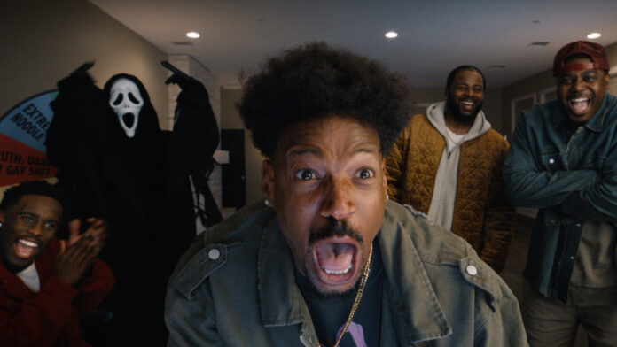 Close-up of a man screaming at the camera, surrounded by friends laughing indoors; a Ghostface-costumed figure stands in the background.