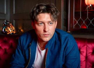 Man in a blue jacket seated on a red tufted sofa in a dim bar, looking to the side.