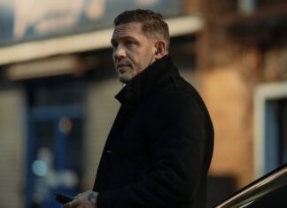 Tom Hardy NÃO foi demitido de ‘Terra da Máfia’, afirma revista Man in a black coat on a city sidewalk, looking to the side and holding a smartphone near a parked car in the background.