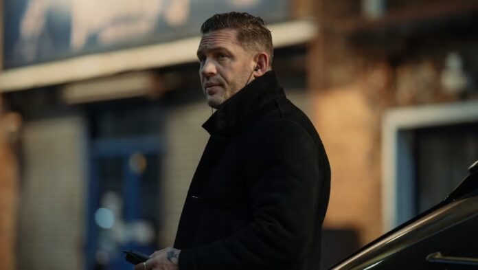 Man in a black coat on a city sidewalk, looking to the side and holding a smartphone near a parked car in the background.