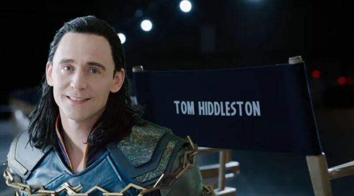 Person in a Loki-inspired costume smiles while seated beside a director's chair with the text 'TOM HIDDLESTON' on the back.