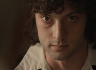 Close-up portrait of a young man with dark curly hair, looking intently at the camera in dim lighting.