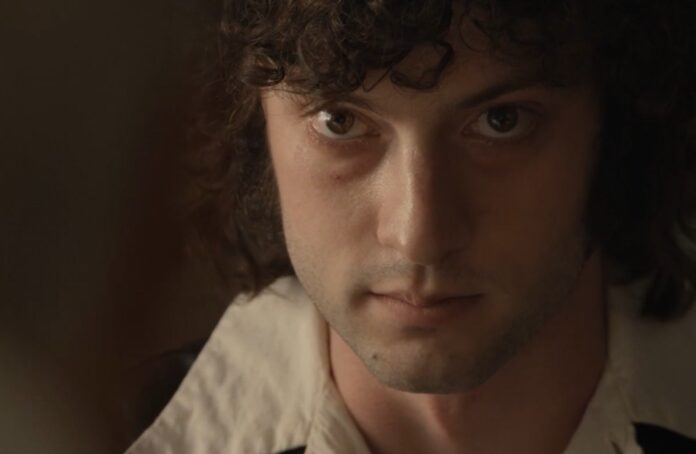 Close-up portrait of a young man with dark curly hair, looking intently at the camera in dim lighting.