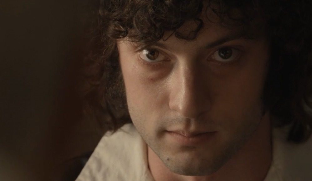 tony a24 Close-up portrait of a young man with dark curly hair, looking intently at the camera in dim lighting.