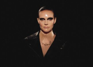 Portrait of a person with slicked-back hair and heavy smoky eye makeup, wearing a black leather jacket in a dark studio.
