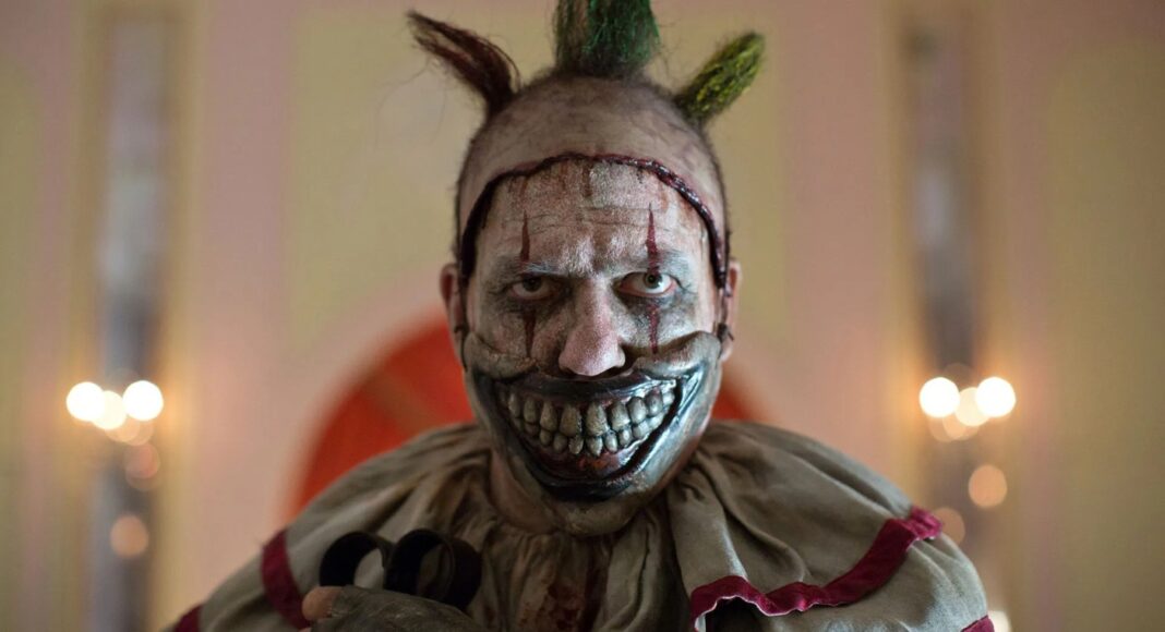 Sinister clown with a wide, bloody-looking grin and metallic silver face paint, green Mohawk, and a tattered beige costume in a dimly lit room.