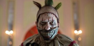 Sinister clown with a wide, bloody-looking grin and metallic silver face paint, green Mohawk, and a tattered beige costume in a dimly lit room.