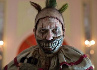 Sinister clown with a wide, bloody-looking grin and metallic silver face paint, green Mohawk, and a tattered beige costume in a dimly lit room.