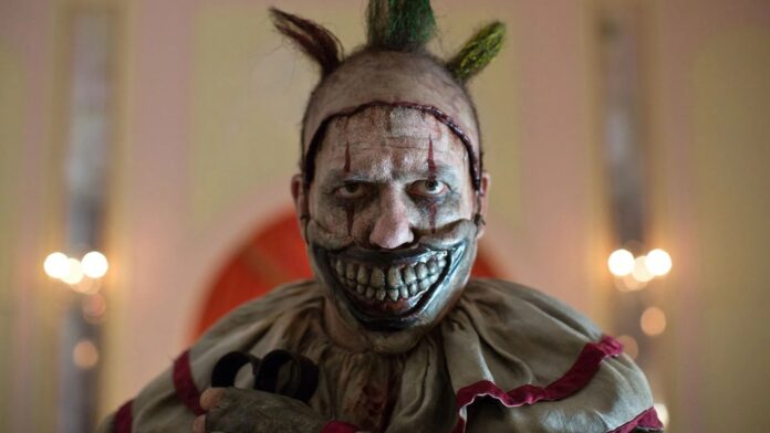 Sinister clown with a wide, bloody-looking grin and metallic silver face paint, green Mohawk, and a tattered beige costume in a dimly lit room.