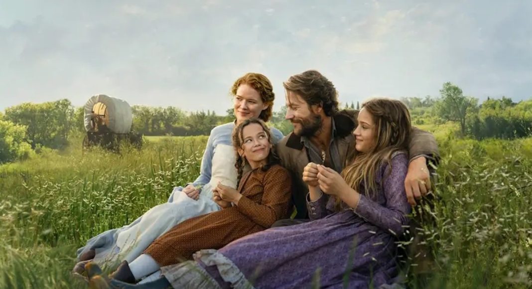 Four people in period costumes sit together in a sunlit grassy field, smiling and relaxing. Nearby, a wagon wheel is visible in the distance.