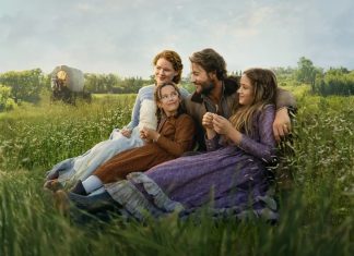 Four people in period costumes sit together in a sunlit grassy field, smiling and relaxing. Nearby, a wagon wheel is visible in the distance.