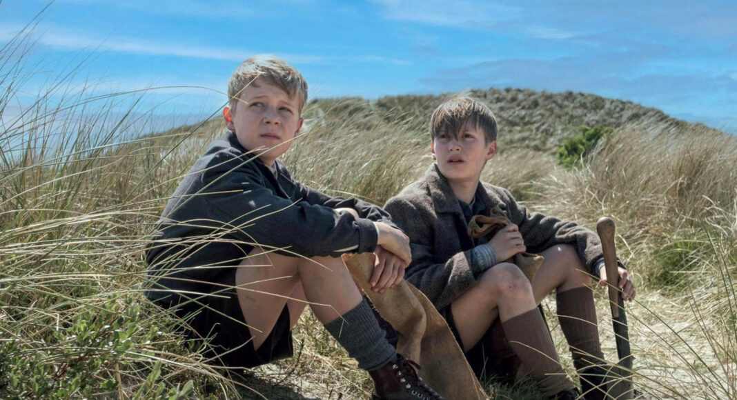 Two boys in vintage clothing sit in sandy dune grass on a sunny day, one holding a shovel.