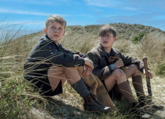Two boys in vintage clothing sit in sandy dune grass on a sunny day, one holding a shovel.