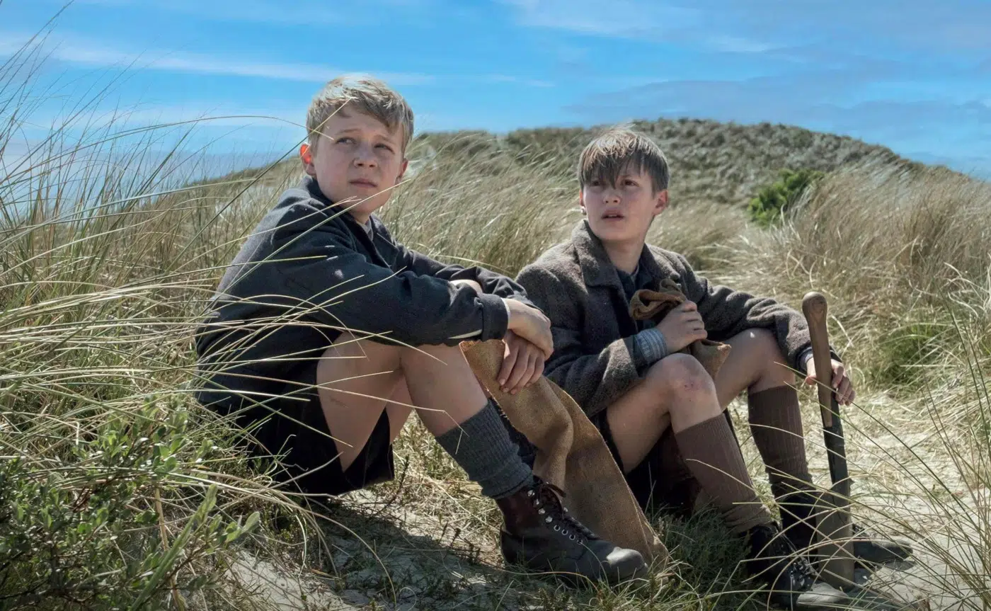Two boys in vintage clothing sit in sandy dune grass on a sunny day, one holding a shovel.