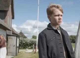 Crítica | Uma Infância Alemã – Drama Aborda as Terríveis Consequências da Guerra para as Crianças Boy in a dark coat stands in a yard, looking off to the right with a serious expression; a woman stands near a house in the background.