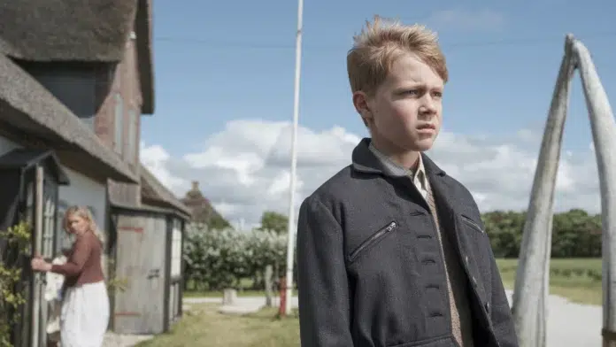 Boy in a dark coat stands in a yard, looking off to the right with a serious expression; a woman stands near a house in the background.