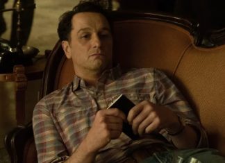 Man in a plaid shirt lounging on an orange sofa, holding a small black wallet or phone in his hands.