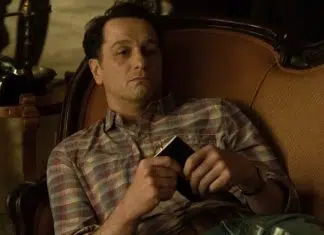 Man in a plaid shirt lounging on an orange sofa, holding a small black wallet or phone in his hands.
