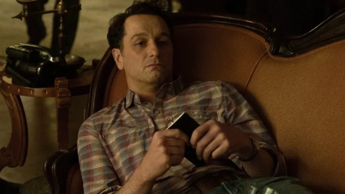 widow's bay 1 Man in a plaid shirt lounging on an orange sofa, holding a small black wallet or phone in his hands.