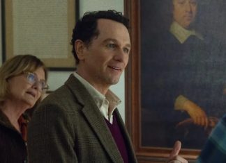 Smiling man in a blazer stands in an art gallery, with a woman in the background and a large portrait on the wall behind them.