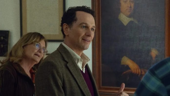 Smiling man in a blazer stands in an art gallery, with a woman in the background and a large portrait on the wall behind them.