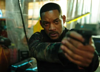 Man in tactical gear aims a handgun directly at the camera in a dim, industrial setting, showing intense focus and readiness to fire.