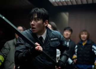 Security guard in a black jacket with a baton, tense expression, in a dim hallway with bystanders behind.