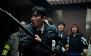 Security guard in a black jacket with a baton, tense expression, in a dim hallway with bystanders behind.