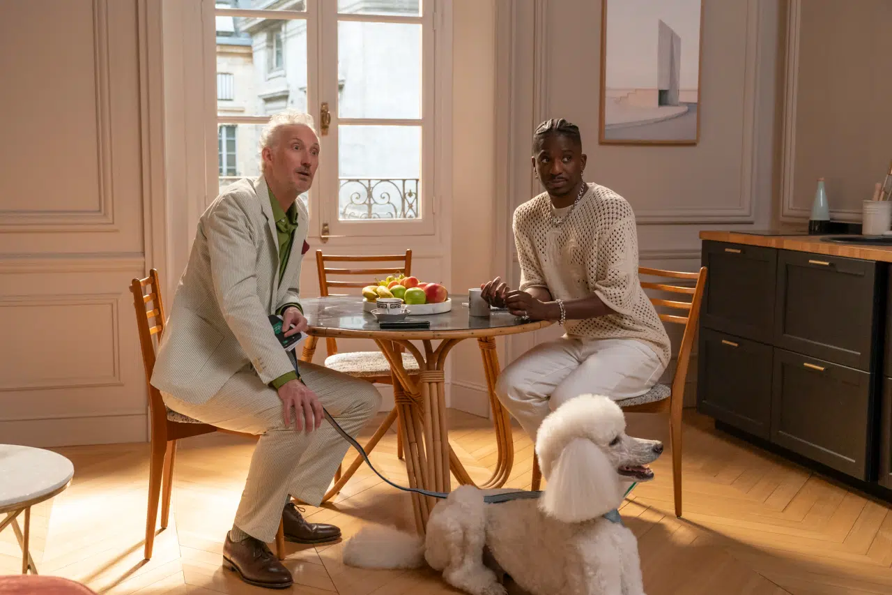 "Two men sitting at a round wooden table in a bright, elegant apartment with a window, fruit bowl, and modern decor. A white poodle on a leash sits on the floor beside them."