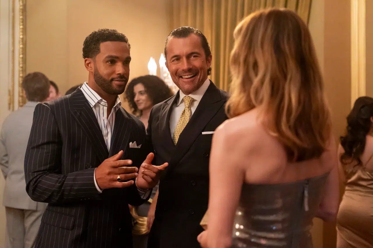 "Two men in suits and a woman in a metallic dress converse at an elegant indoor party, surrounded by other formally dressed guests in a well-lit, upscale setting with gold accents and curtains."