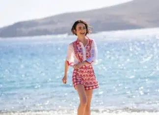 "A young woman in a patterned shirt and shorts smiles while walking along a sunny beach, with sparkling blue water and distant hills in the background."